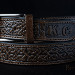 May include: A brown leather belt featuring an embossed Celtic knot pattern and the letters KC stamped on the strap. The belt includes a silver-toned metal buckle and a matching leather keeper loop, presented against a dark background with the text Brown.