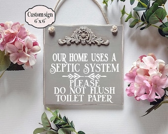May include: A grey wooden sign with the text "OUR HOME USES A SEPTIC SYSTEM PLEASE DO NOT FLUSH TOILET PAPER." The sign is 15.24 cm x 15.24 cm and has a decorative floral design. It hangs from a rope and is surrounded by pink flowers and greenery.