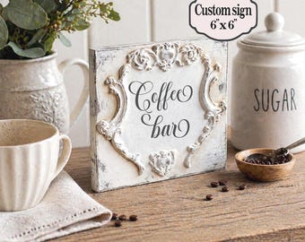 May include: A distressed white wooden sign with the words "Coffee bar" in elegant script. The sign is 6 x 6 inches and features ornate, raised detailing. A coffee cup, sugar jar, and decorative elements complete the coffee-themed display.