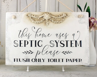May include: White wooden sign with a distressed finish and a decorative floral design. The sign reads "This home uses a septic system please flush only toilet paper".