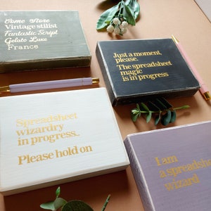 May include: Four wooden signs with gold lettering. The signs have different backgrounds and sayings. The first sign is green and says "Come Store Vintage stilist Fantastic Script Gelato Luxe France". The second sign is black and says "Just a moment, please. The spreadsheet magic is in progress". The third sign is white and says "Spreadsheet wizardry in progress. Please hold on". The fourth sign is purple and says "I am a spreadsheet wizard".