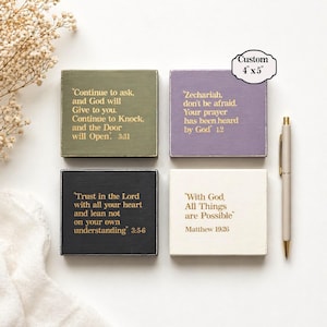 May include: Four square wooden blocks with inspirational quotes in gold lettering. The blocks are olive green, lavender, black, and cream, each 10 x 13 cm. A gold-tipped pen rests beside the blocks. The text includes Bible verses.
