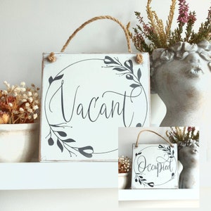 May include: Two wooden signs with a white background and black floral designs. The top sign says "Vacant" and the bottom sign says "Occupied".