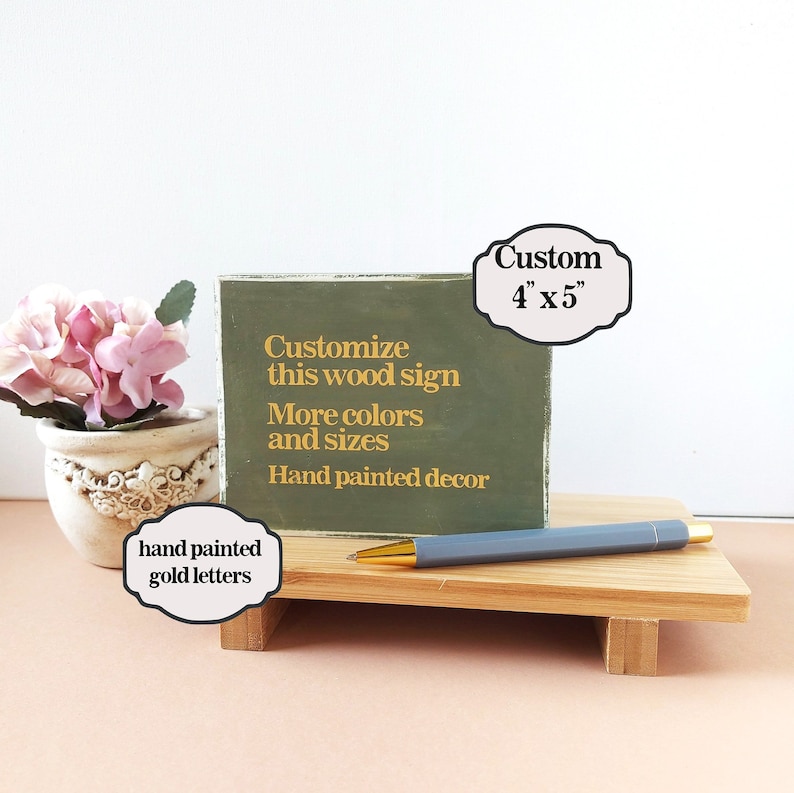 May include: A green wooden sign with gold lettering that reads "Customize this wood sign More colors and sizes Hand painted decor". The sign is 4 inches by 5 inches and is sitting on a wooden stand. The sign is hand painted with gold letters.