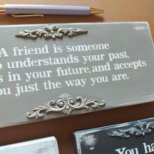 May include: A gray wooden sign with a quote about friendship in white lettering, framed by ornate silver-colored embellishments. A pen with a gold tip and a lavender barrel rests above the sign. Other signs are visible.