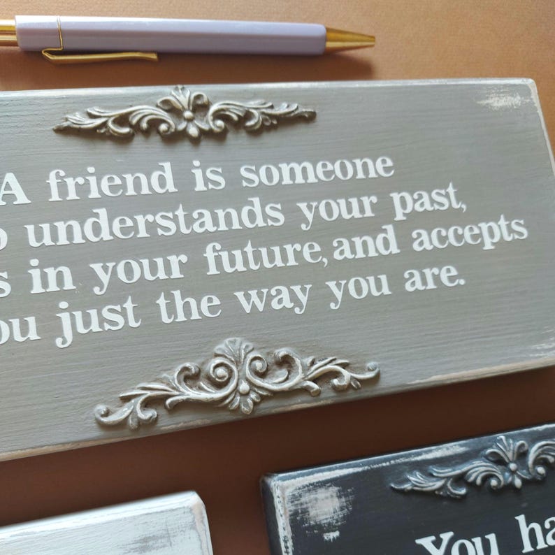 May include: A gray wooden sign with a quote about friendship in white lettering, framed by ornate silver-colored embellishments. A pen with a gold tip and a lavender barrel rests above the sign. Other signs are visible.