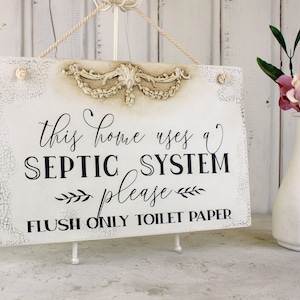 May include: White wooden sign with black text that reads "This home uses a septic system please flush only toilet paper". The sign has a decorative, ornate, white, raised design at the top.