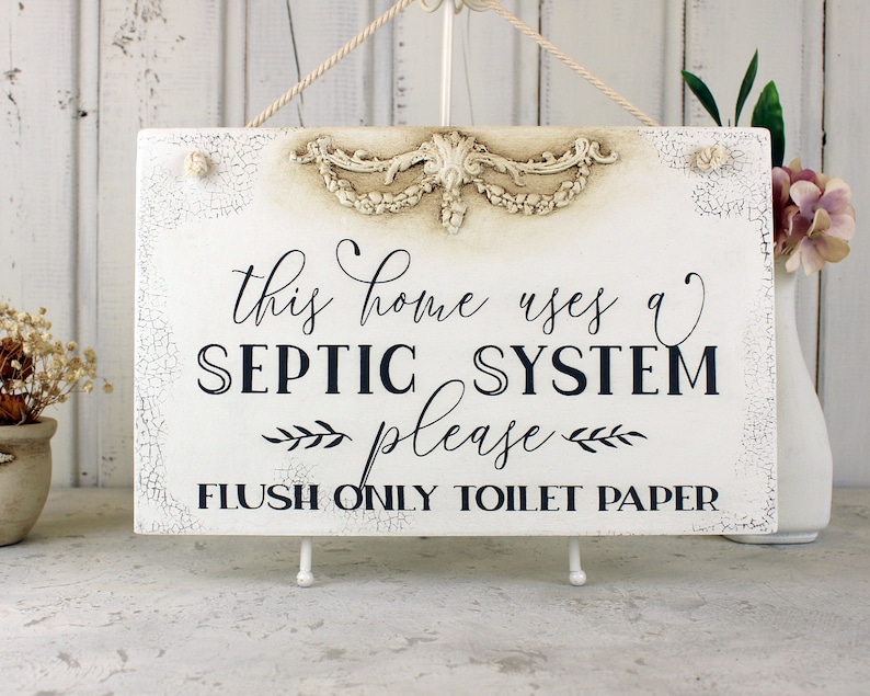 May include: White wooden sign with a distressed finish and a decorative floral design. The sign reads "This home uses a septic system please flush only toilet paper".