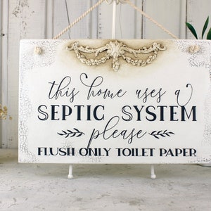 May include: White sign with distressed paint and a decorative floral design at the top. The sign reads "This home uses a septic system please flush only toilet paper".