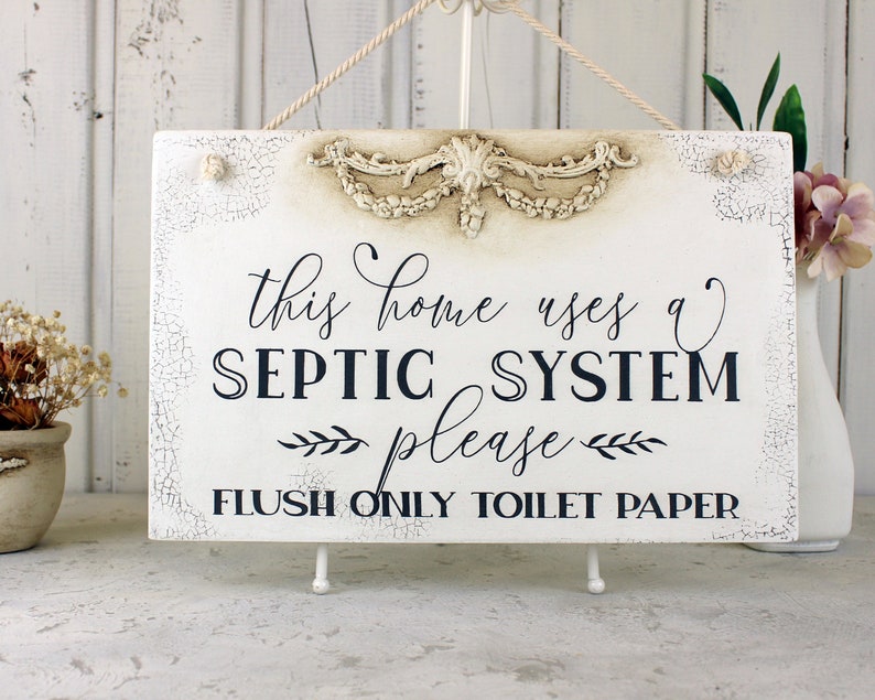 May include: White sign with distressed paint and a decorative floral design at the top. The sign reads "This home uses a septic system please flush only toilet paper".
