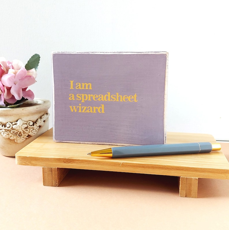 May include: A purple wooden sign with the text "I am a spreadsheet wizard" in gold lettering. The sign is sitting on a wooden stand with a gray pen on top.