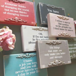 May include: A collection of decorative wooden signs in various colors, each featuring inspirational quotes in white text. The signs have ornate, decorative accents and are arranged against a green background. The quotes include phrases about self-improvement and life choices.