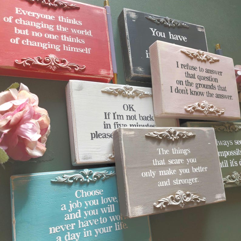 May include: A collection of decorative wooden signs in various colors, each featuring inspirational quotes in white text. The signs have ornate, decorative accents and are arranged against a green background. The quotes include phrases about self-improvement and life choices.