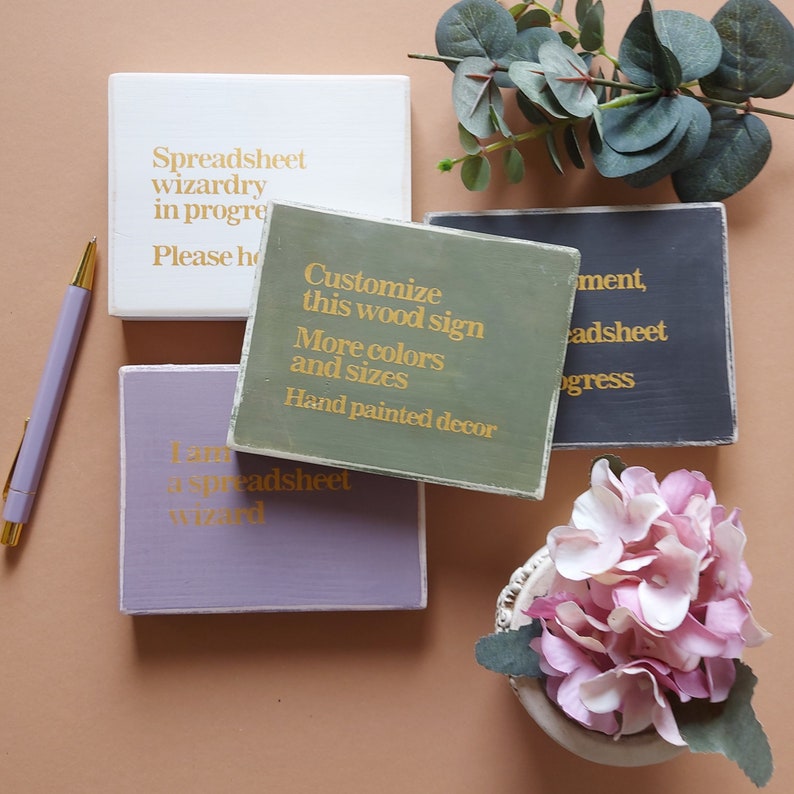 May include: Four wooden signs with gold lettering. The signs have different colors and sayings. The signs read: "Spreadsheet wizardry in progress, Please hold", "Customize this wood sign, More colors and sizes, Hand painted decor", "I am a spreadsheet wizard", and "ment, adsheet progress".