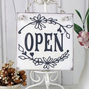 Peut inclure: Une pancarte en bois blanc avec un motif floral noir et le mot "OUVERT" en lettres noires.