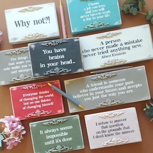 Op de afbeelding: Een verzameling decoratieve houten borden met inspirerende citaten in verschillende kleuren, waaronder wit, grijs, rood en groen. Elk bord heeft sierlijke decoraties en tekst. Pennen liggen verspreid rond de borden.