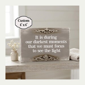 May include: A gray wooden sign, 4" x 6", with the text "It is during our darkest moments that we must focus to see the light." The sign has ornate silver accents and is displayed on a light-colored surface.
