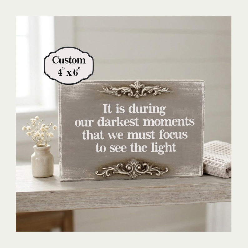 May include: A gray wooden sign, 4" x 6", with the text "It is during our darkest moments that we must focus to see the light." The sign has ornate silver accents and is displayed on a light-colored surface.