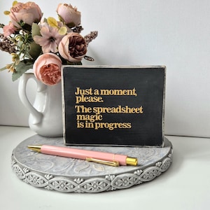 May include: A black wooden sign with gold text: "Just a moment, please. The spreadsheet magic is in progress." A pink pen with gold details sits on a decorative grey stand. A white jug holds pink and peach flowers.