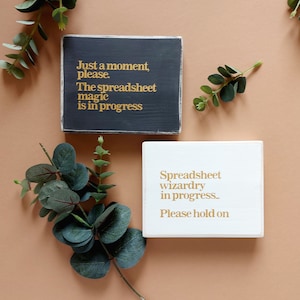 May include: Two wooden signs with gold lettering. The first sign is black and says "Just a moment, please. The spreadsheet magic is in progress". The second sign is white and says "Spreadsheet wizardry in progress. Please hold on".