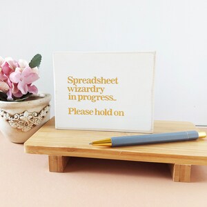 May include: White sign with gold text that reads "Spreadsheet wizardry in progress. Please hold on". The sign is sitting on a wooden stand with a gray pen on top.