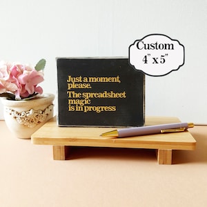 May include: A black wooden sign with gold lettering that reads "Just a moment, please. The spreadsheet magic is in progress". The sign is sitting on a wooden stand with a pen on top of the stand. The sign is 4 inches by 5 inches.