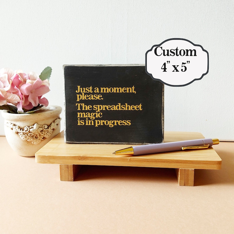 May include: A black wooden sign with gold lettering that reads "Just a moment, please. The spreadsheet magic is in progress". The sign is sitting on a wooden stand with a pen on top of the stand. The sign is 4 inches by 5 inches.