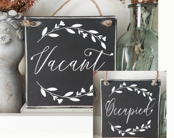 May include: Two black wooden signs with white floral wreaths and the words "Vacant" and "Occupied" written in white cursive.