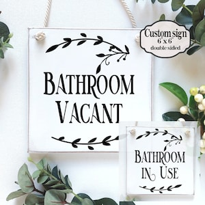 May include: A white wooden sign with black text and floral accents. The sign reads "Bathroom Vacant" on one side and "Bathroom In Use" on the other side. The sign is 6 inches by 6 inches and is double-sided.