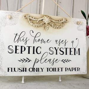 May include: White sign with distressed paint and a decorative floral design at the top. The sign reads "This home uses a septic system please flush only toilet paper".