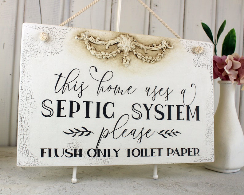 May include: White sign with distressed paint and a decorative floral design at the top. The sign reads "This home uses a septic system please flush only toilet paper".