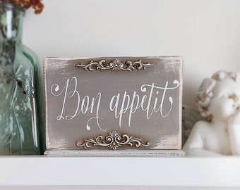 May include: A rustic wooden sign with a distressed grey finish and white lettering that reads "Bon appetit". The sign has decorative metal accents around the edges.