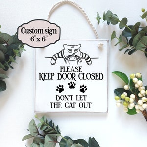 Può includere: Un cartello di legno bianco con un'illustrazione in bianco e nero di un gatto che sporge dalla parte superiore. Il cartello dice "Si prega di tenere la porta chiusa, non lasciare che il gatto esca".