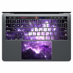 May include: A purple and white galaxy themed keyboard skin for a Macbook Pro laptop. The skin covers the keys and the trackpad.