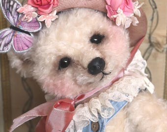 May include: A cream-colored teddy bear wearing a light blue denim overall with a small silver wing pin. The bear has a pink straw hat adorned with pink fabric roses and a purple butterfly, and a white lace collar with a pink velvet ribbon tie.