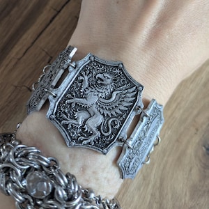 May include: A silver-toned cuff bracelet featuring a central shield-shaped plaque with an embossed griffin design, connected by metal links to side panels. Below it, a separate silver-toned chainmail-style bracelet with a central round stone.