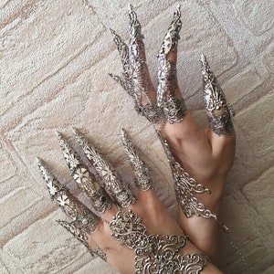 May include: A pair of silver metal finger rings with intricate designs. The rings are connected by a chain and have long, pointed tips.