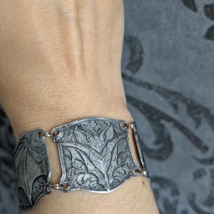 Può includere: Un bracciale in metallo color argento composto da una serie di maglie rettangolari, ciascuna con un motivo a pipistrello dettagliato e intricati disegni a volute. Le maglie sono collegate da piccoli anelli metallici per un design flessibile.