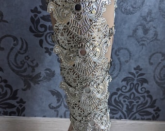 May include: A silver-toned metal leg cuff featuring an intricate filigree pattern with repeating ornate scrollwork and floral motifs. The structure consists of multiple connected panels that wrap around the lower leg, secured by small metal side clasps.