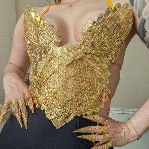 May include: A gold-colored, ornate, decorative, metal top with a detailed floral design. The top is worn over the chest and has a halter-style strap.