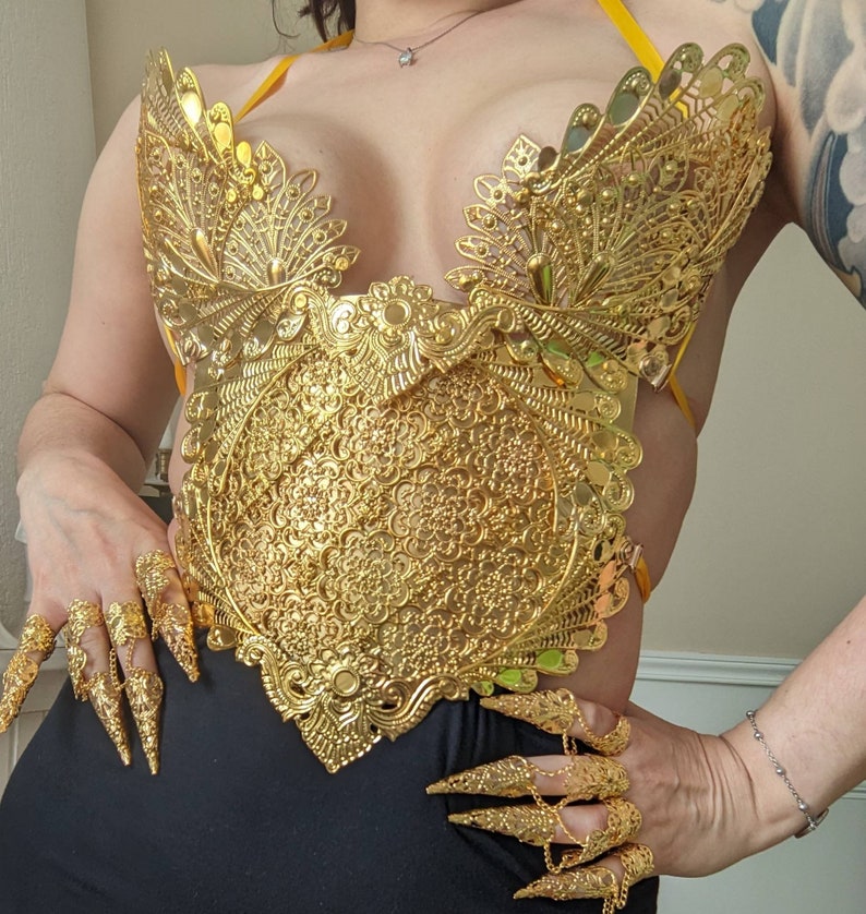 May include: A gold-colored, ornate, decorative, metal top with a detailed floral design. The top is worn over the chest and has a halter-style strap.