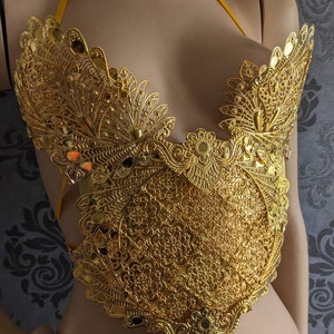May include: A gold-colored, ornate, decorative bra top with a delicate, intricate design. The top is made of metal and has a floral pattern.
