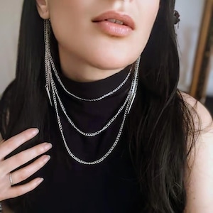 Peut inclure: Un collier argenté à plusieurs rangs avec trois chaînes. Le collier a un pendentif en forme de pointe aux extrémités des deux chaînes supérieures. Le modèle porte un col roulé noir.