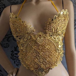 May include: A gold-colored, intricate, and ornate metal bra top with a delicate floral design. The bra top has a halter style with a yellow strap.