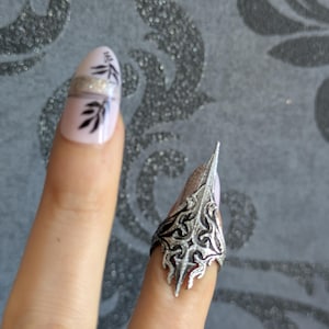 May include: A silver-colored finger armor ring with an ornate, gothic design. The ring is worn on a finger with a light pink nail polish with black leaf designs and a silver glitter band.
