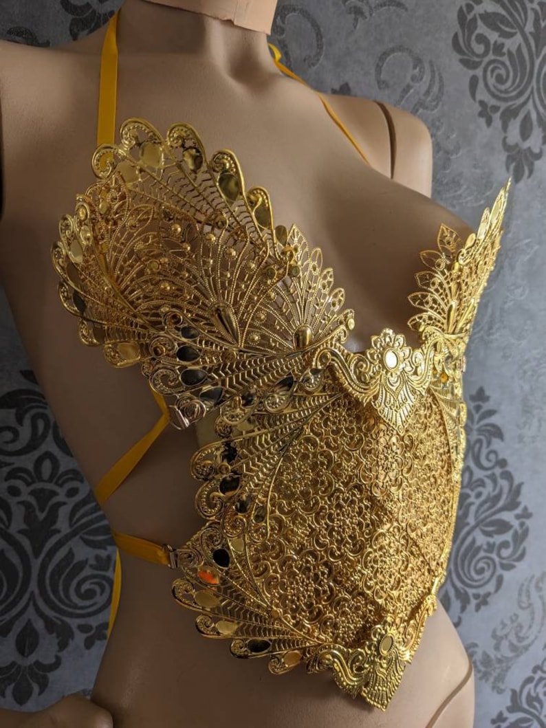 May include: A gold-colored, intricate, and detailed, decorative,  halter-style top with a delicate, lacy design. The top is made of metal and has a  peacock-like pattern.