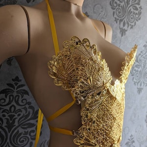 May include: A gold-colored, ornate, decorative bra with a yellow ribbon halter strap. The bra is made of intricate, filigree-like metalwork.
