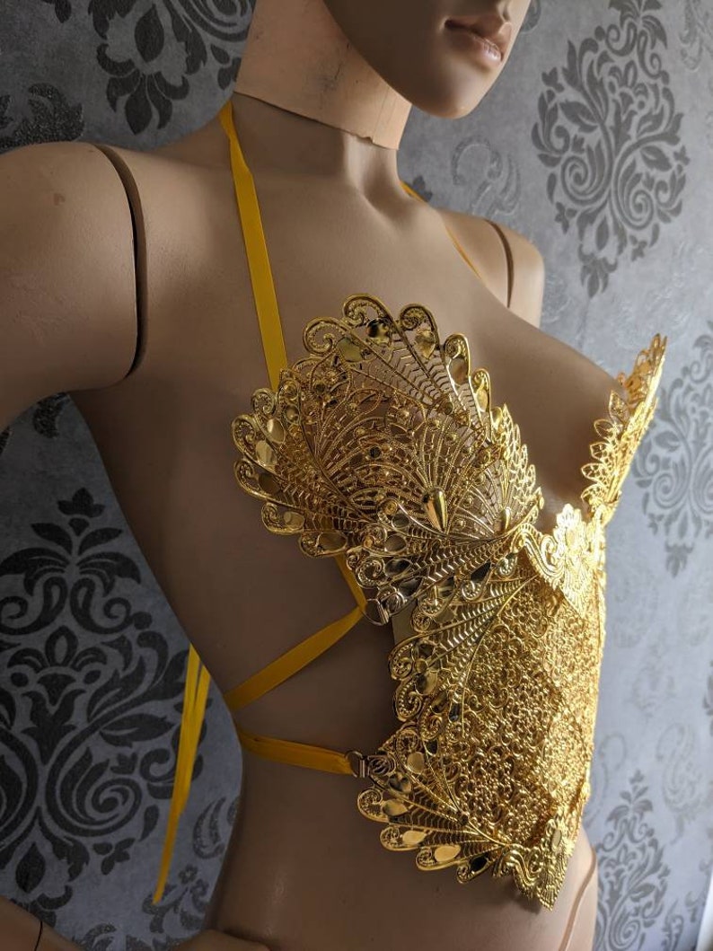 May include: A gold-colored, ornate, decorative bra with a yellow ribbon halter strap. The bra is made of intricate, filigree-like metalwork.