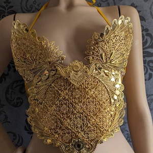 May include: A gold-colored, ornate, decorative top with a delicate, intricate design. The top is made of metal and has a circular center piece with a floral pattern. The top is on a mannequin.
