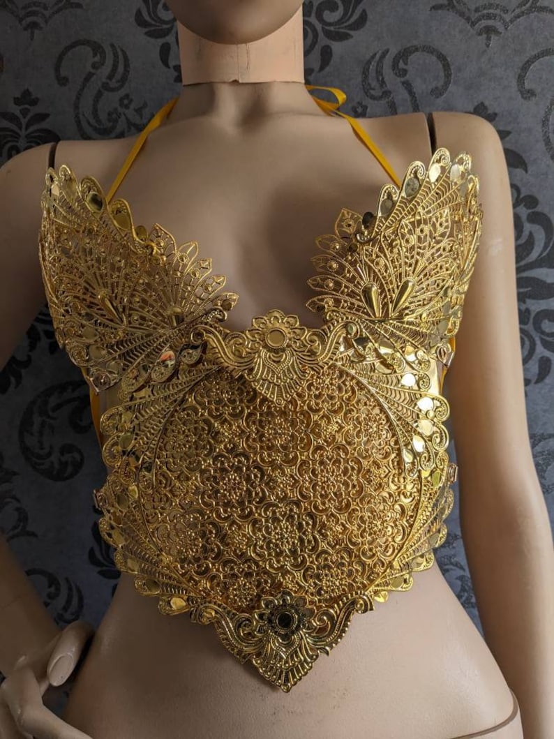 May include: A gold-colored, ornate, decorative top with a delicate, intricate design. The top is made of metal and has a circular center piece with a floral pattern. The top is on a mannequin.